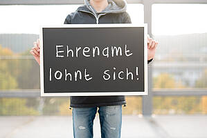 Eine Person hält ein Schild hoch, auf dem steht: Ehrenamt lohnt sich!